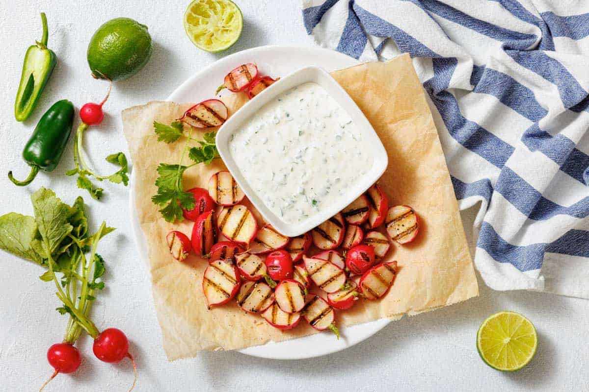 A plate with grilled radishes, a bowl of creamy dipping sauce, lime halves, jalape&ntilde;o, and a striped cloth on a white surface.