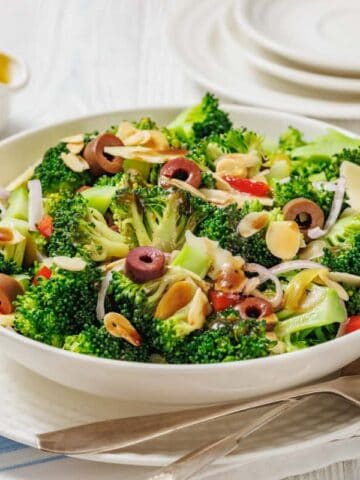 A bowl of Italian broccoli salad with sliced olives, almonds, onions, and red peppers, served on a white plate with a fork and napkin beside it.