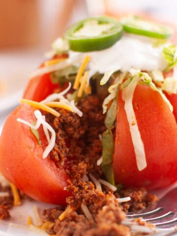 A sliced tomato stuffed with seasoned ground meat, shredded cheese, lettuce, sour cream, and jalapeño slices is served on a white plate with a fork.