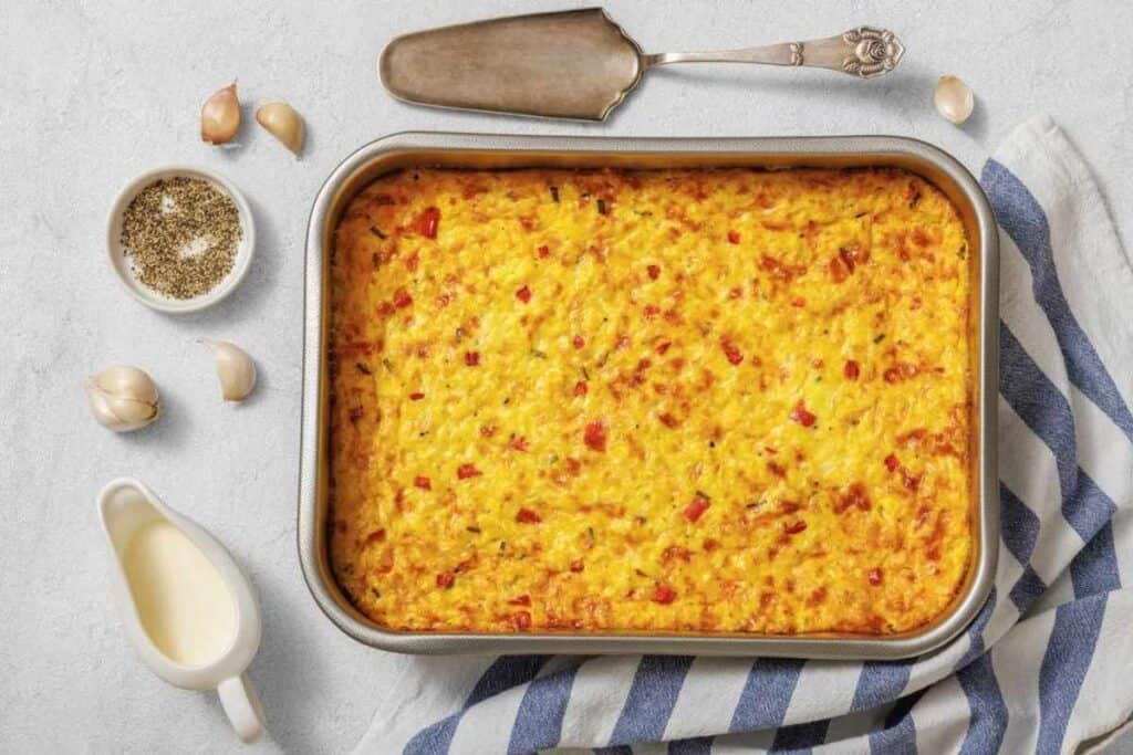 A baked casserole with a golden crust in a metal pan, placed on a striped towel, with garlic, black pepper, cream, and a serving utensil nearby.