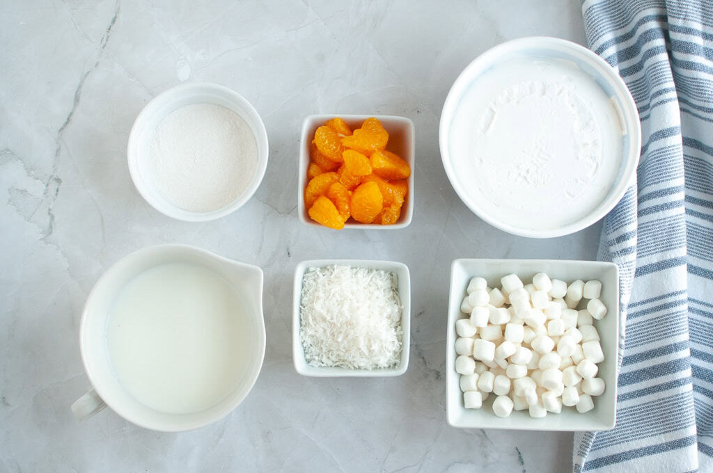 Overhead shot of ingredients for Orange Fluff Salad: whipped topping, mandarin oranges, marshmallows, coconut flakes, and powdered sugar.