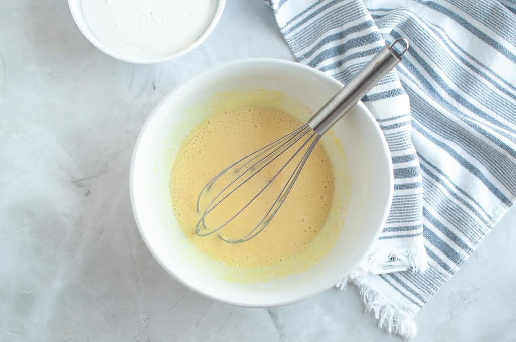 Overhead shot of a pale yellow lemon curd mixture being whisked in a white bowl.