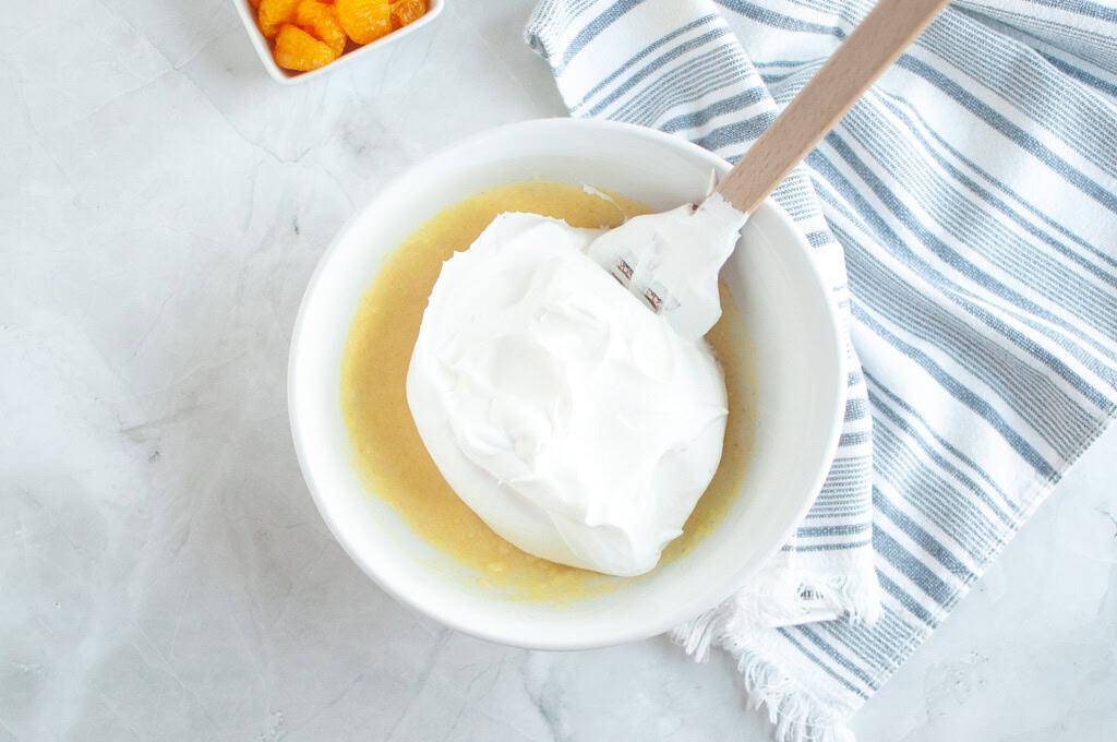 Overhead shot of white whipped topping being gently folded into a pale yellow mixture in a white bowl with a spatula.