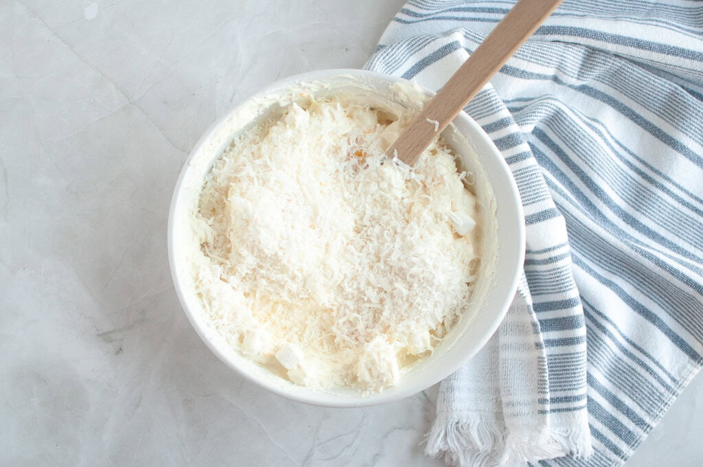 Overhead shot of shredded coconut being folded into a creamy, light-colored mixture in a white bowl with a spatula.