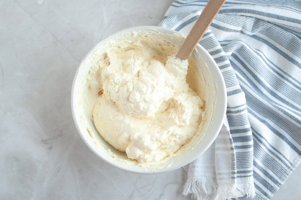 Overhead shot of a white bowl filled with creamy, light-colored Orange Fluff Salad being mixed with a spatula.