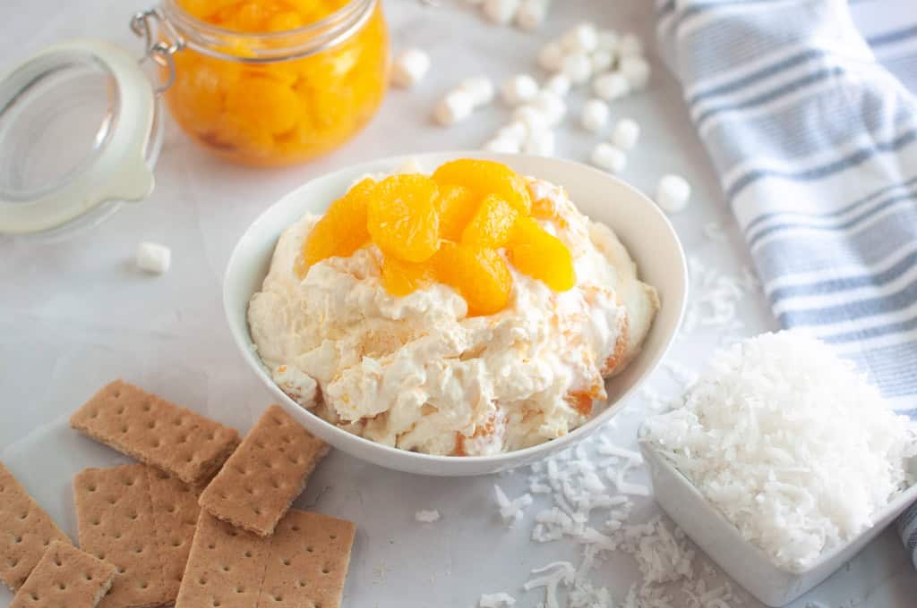 Overhead shot of a white bowl filled with creamy Orange Fluff Salad topped with mandarin oranges, surrounded by graham crackers and marshmallows.