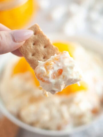 A hand holds a cracker topped with Orange Fluff Salad Dip over a bowl of the dip.
