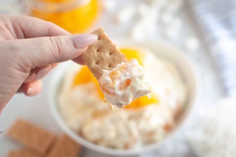 A hand holds a cracker topped with Orange Fluff Salad Dip over a bowl of the dip.