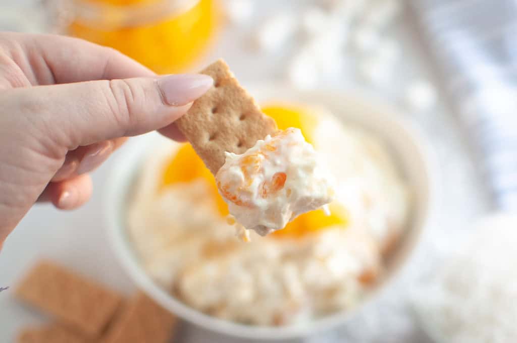A hand holds a cracker topped with Orange Fluff Salad Dip over a bowl of the dip.