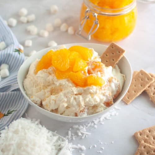 Overhead shot of a white bowl filled with creamy Orange Fluff Salad, topped with mandarin oranges and served with graham crackers.