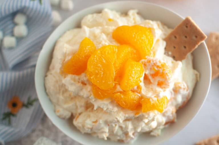 Overhead shot of a white bowl filled with creamy orange fluff salad, topped with mandarin oranges and served with graham crackers.