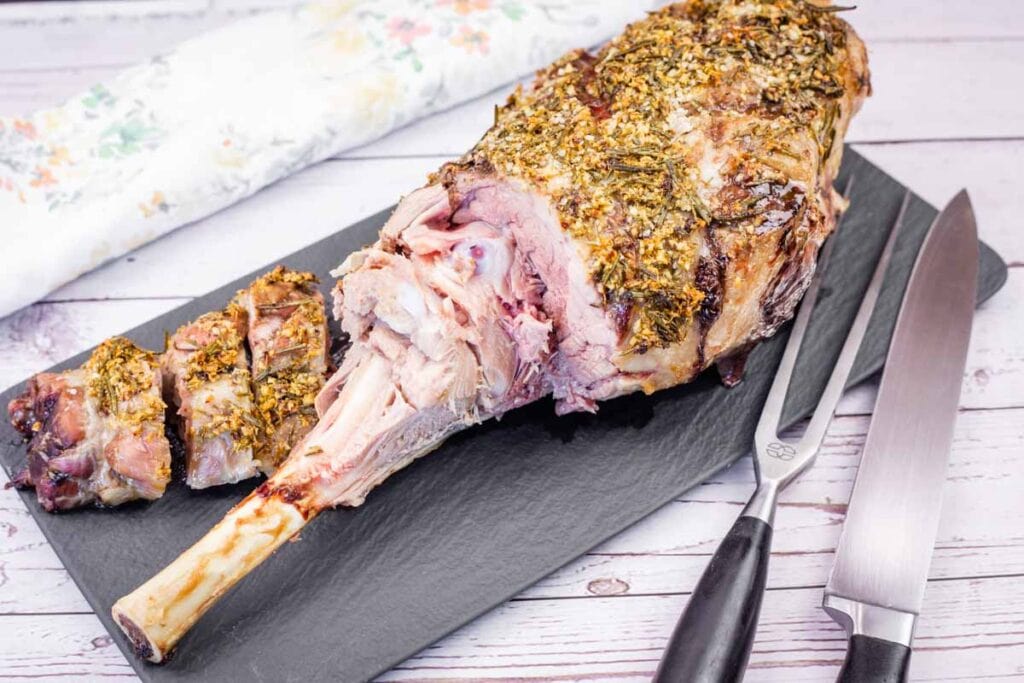 Roast leg of lamb with herb crust, partially sliced, displayed on a slate board with a carving fork and knife beside it.