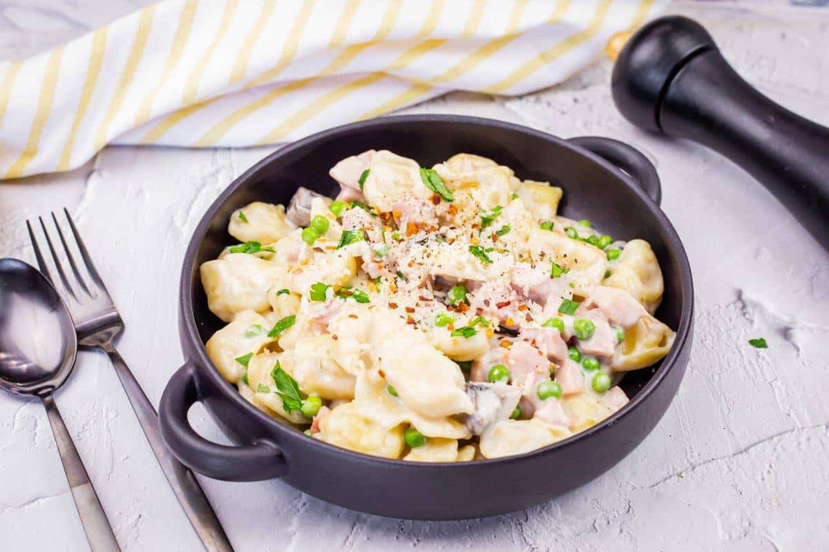 A black bowl of Easy Skillet Cheese Tortellini with Ham & Peas garnished with grated cheese and herbs.