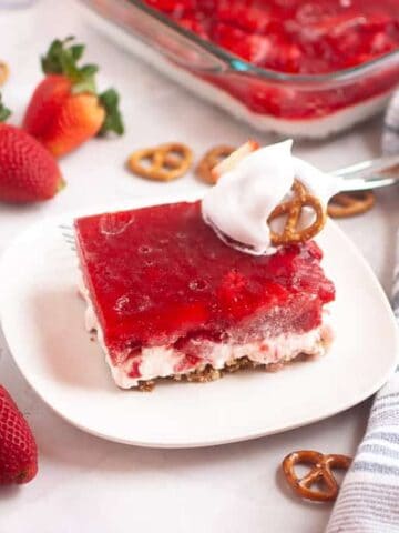 A slice of Strawberry Pretzel Salad topped with whipped cream and a pretzel sits on a white plate.
