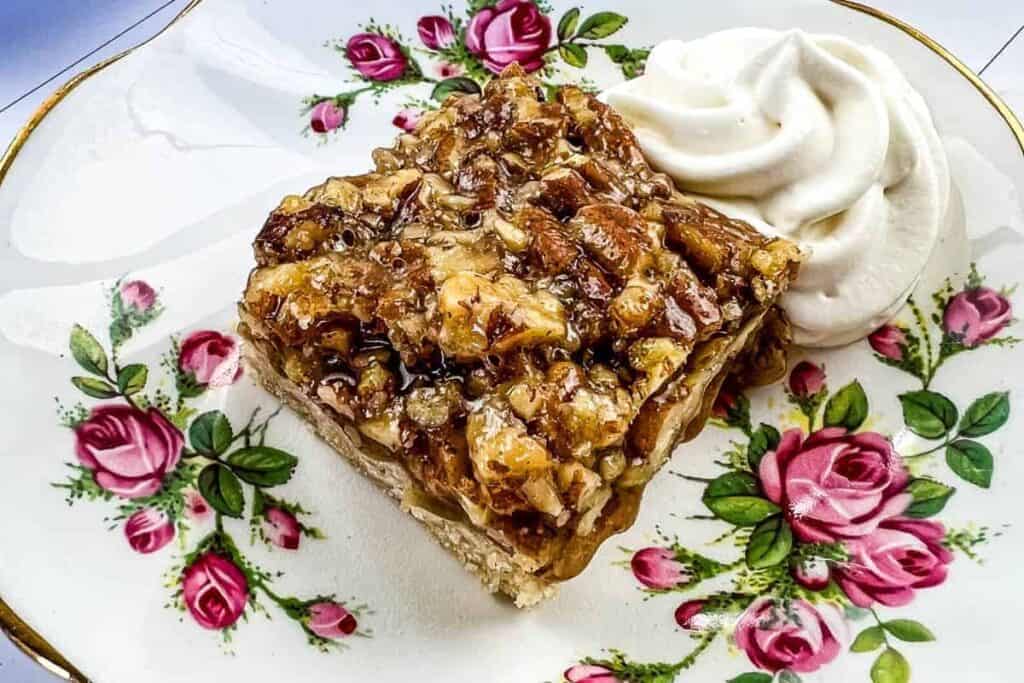 A square pecan dessert bar sits on a floral plate beside a swirl of whipped cream.