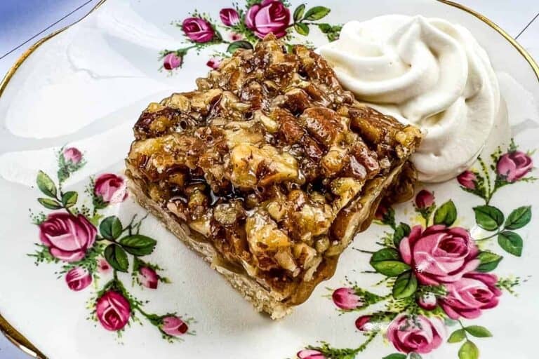 A square pecan dessert bar sits on a floral plate beside a swirl of whipped cream.