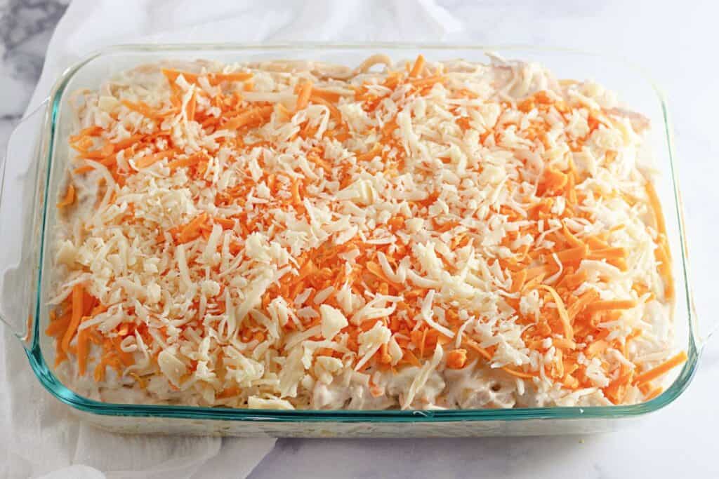 A glass baking dish filled with an unbaked casserole, topped with shredded cheese and grated carrots, placed on a white surface.
