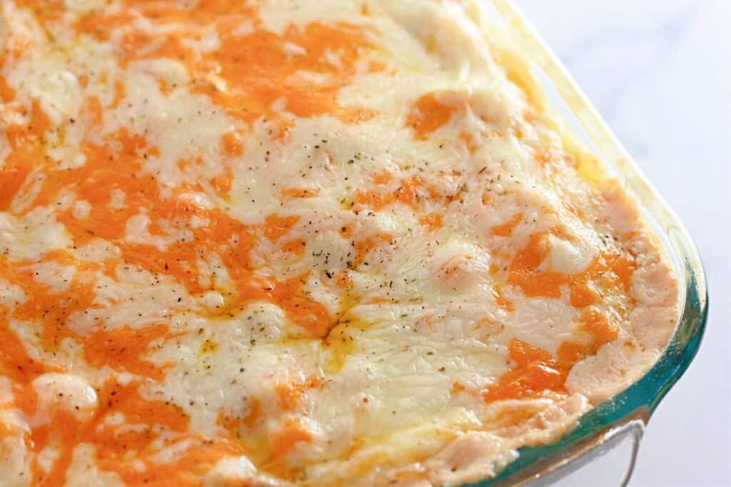A close-up of a baked casserole dish topped with melted white and orange cheese, lightly seasoned with black pepper.