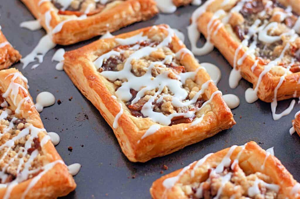 A close-up of several rectangular Dutch Apple Danishes with flaky pastry, apple filling, crumb topping, and white icing drizzle, on a dark baking sheet.