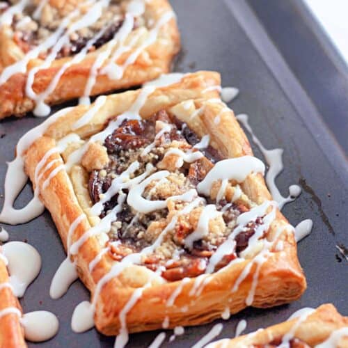 A close-up of three rectangular Dutch Apple Danishes with flaky pastry, apple filling, crumb topping, and white icing drizzle, on a dark baking sheet.