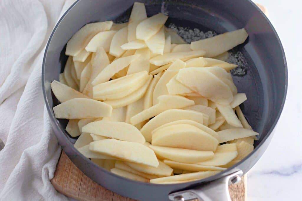 Thinly sliced raw apples in a gray skillet, ready for cooking, placed on a wooden board with a white kitchen towel nearby.