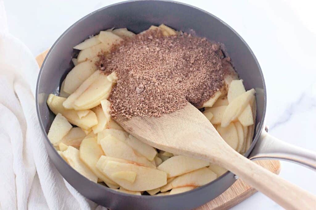Sliced apples in a gray skillet topped with a generous amount of cinnamon sugar mixture, being stirred with a wooden spatula.