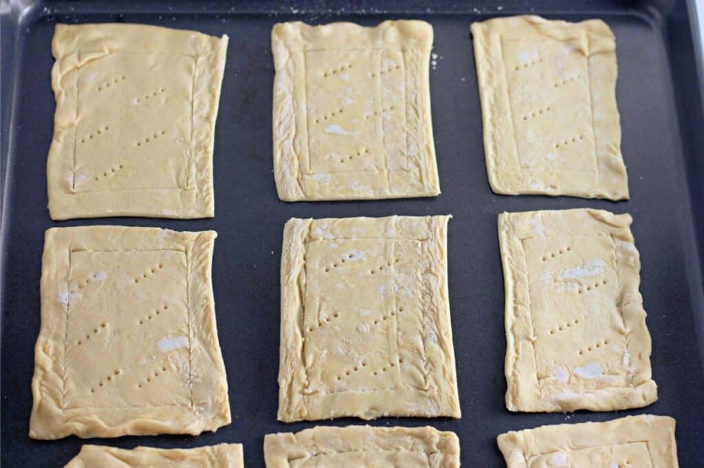 Six rectangular sheets of light tan, uncooked pastry dough with pricked surfaces are neatly arranged on a dark metal baking sheet.