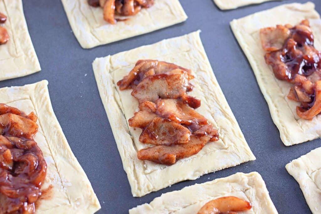 Rectangular pieces of uncooked pastry dough are arranged on a dark baking sheet, each topped with a glossy, reddish-brown apple filling.