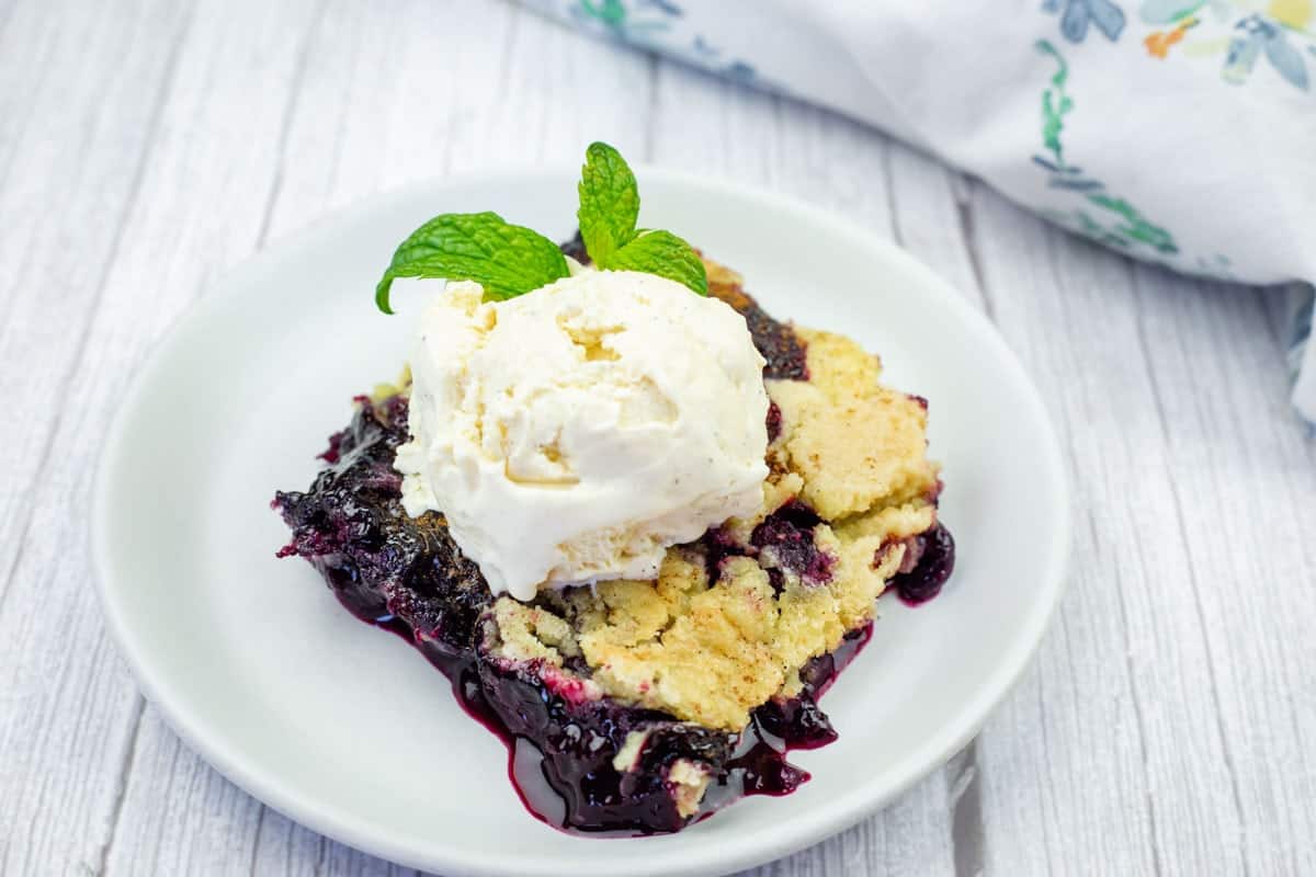 A serving of blueberry dump cake topped with a scoop of vanilla ice cream and a sprig of mint on a white plate.