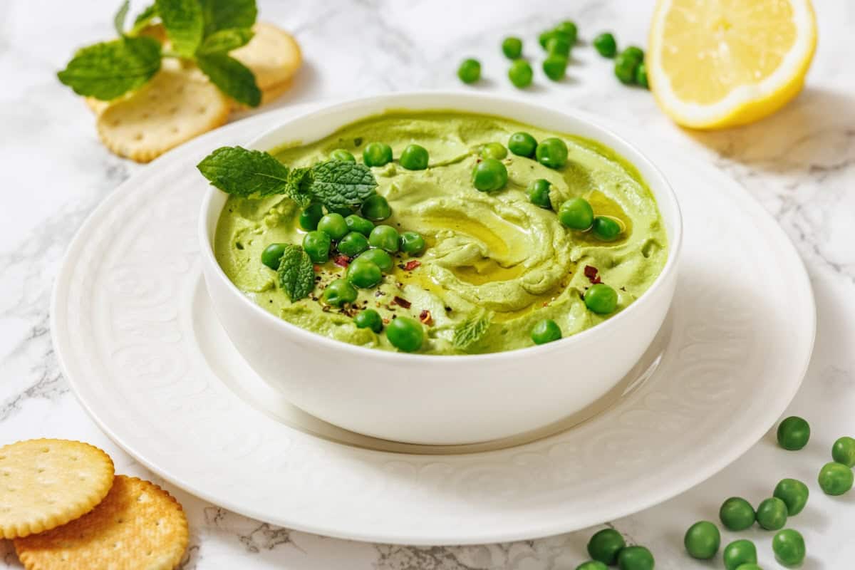 A white bowl of vibrant green Green Pea Hummus, garnished with peas and mint, served with crackers and a lemon wedge.