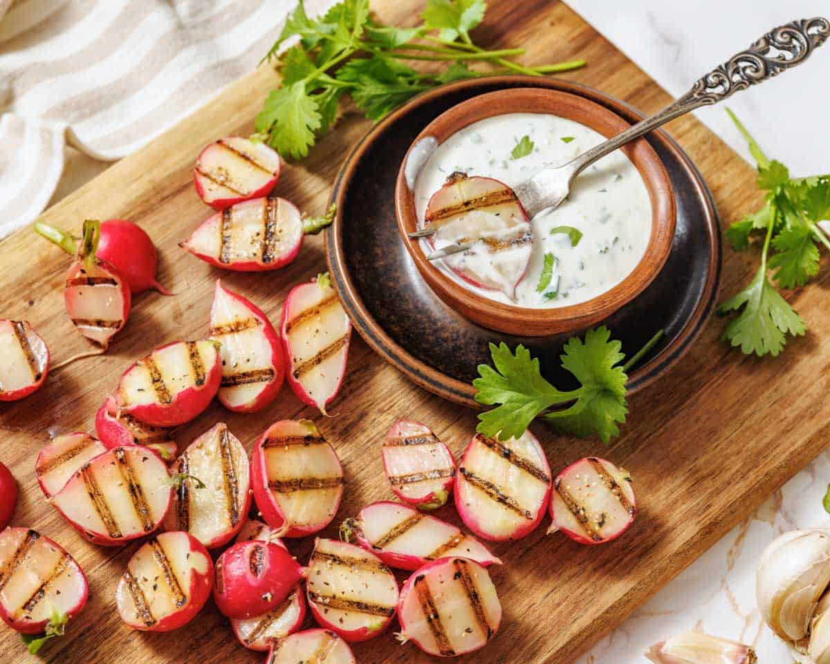 Grilled radish halves with char marks served on a rustic wooden board, accompanied by a small bowl of creamy jalape&ntilde;o dip garnished with cilantro and a decorative spoon, surrounded by fresh herbs and garlic cloves.