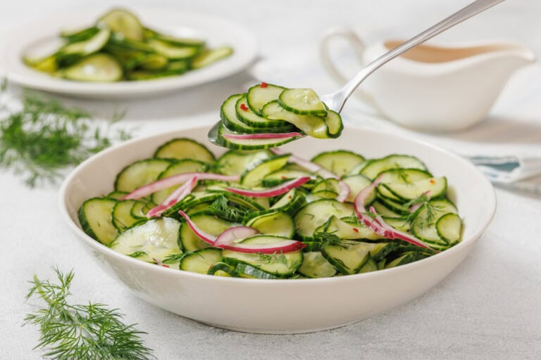 A bowl of cucumber salad with red onion slices and dill, with a spoon lifting a portion. A plate of salad and a sauce boat are in the background.