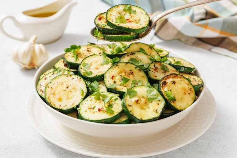 A white bowl filled with sliced, seasoned, and roasted zucchini garnished with fresh herbs; a spoon is lifting some zucchini slices. A garlic bulb and sauce pitcher are in the background.