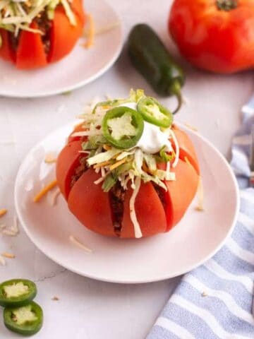 A tomato cut into wedges and filled with taco ingredients, topped with shredded cheese, sour cream, and sliced jalape&ntilde;os on a white plate. Nearby are cheese shreds, jalape&ntilde;o slices, and a fork.