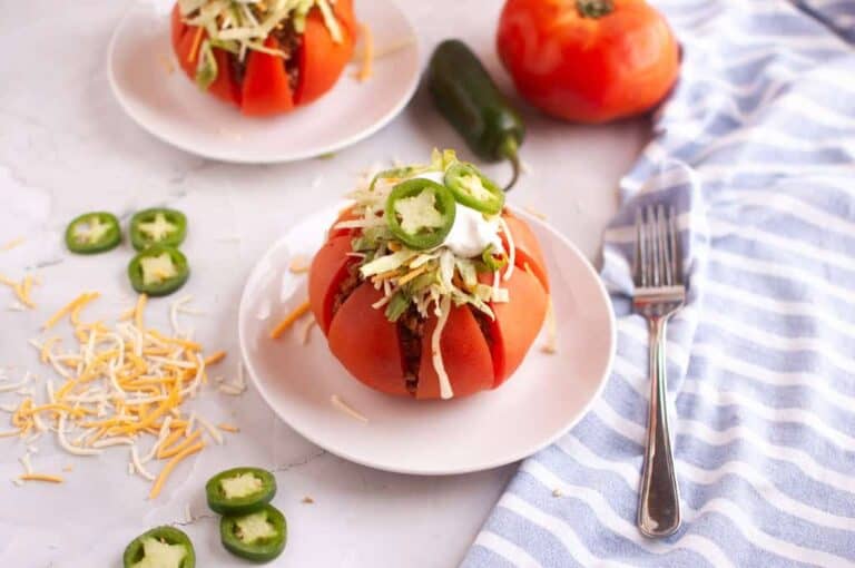 A tomato cut into wedges and filled with taco ingredients, topped with shredded cheese, sour cream, and sliced jalapeños on a white plate. Nearby are cheese shreds, jalapeño slices, and a fork.