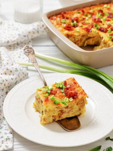 A square slice of baked casserole with cheese and green onions on a white plate, with the rest of the casserole in a baking dish in the background.