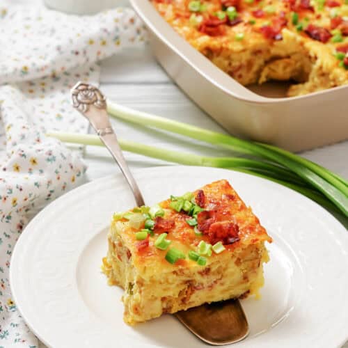 A square slice of baked casserole with cheese and green onions on a white plate, with the rest of the casserole in a baking dish in the background.