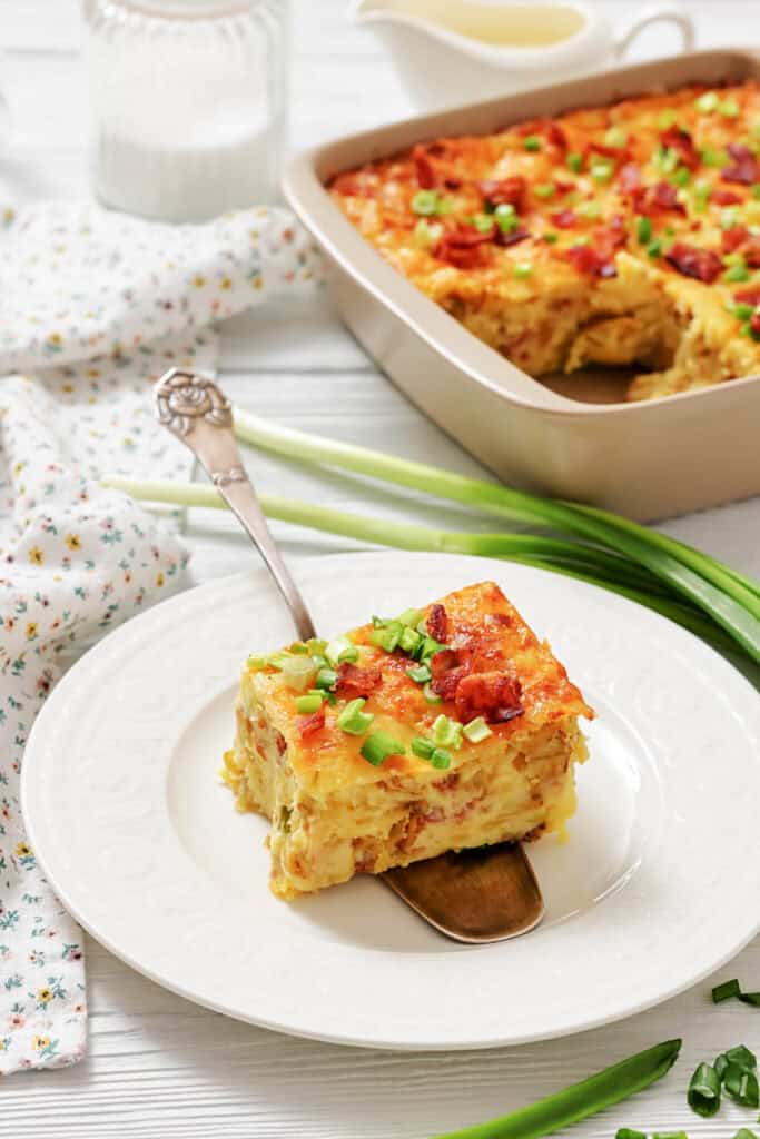A square slice of baked casserole with cheese and green onions on a white plate, with the rest of the casserole in a baking dish in the background.