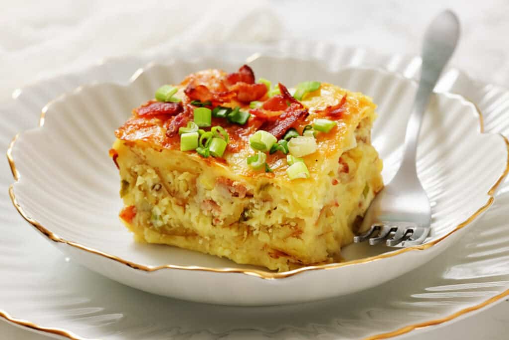 A slice of breakfast casserole topped with chopped green onions and bacon sits on a white plate with a fork.