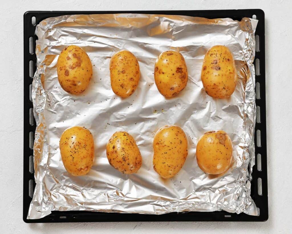 Eight seasoned whole potatoes are arranged on a foil-lined baking tray, ready to be cooked.