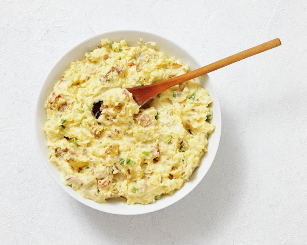A bowl of potato salad with chopped herbs, mixed with mayonnaise, and a wooden spoon resting inside.