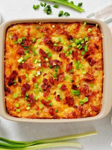 A twice-baked potato casserole topped with chopped green onions and bacon bits sits on a table alongside green onions, pepper, salt, cream, and a serving utensil.