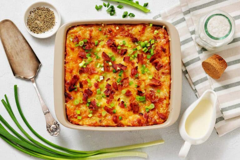 A twice-baked potato casserole topped with chopped green onions and bacon bits sits on a table alongside green onions, pepper, salt, cream, and a serving utensil.