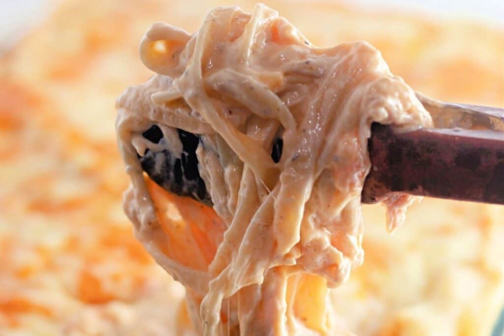 Close-up of metal tongs lifting a serving of melted, stringy cheese from a cheesy baked dish.