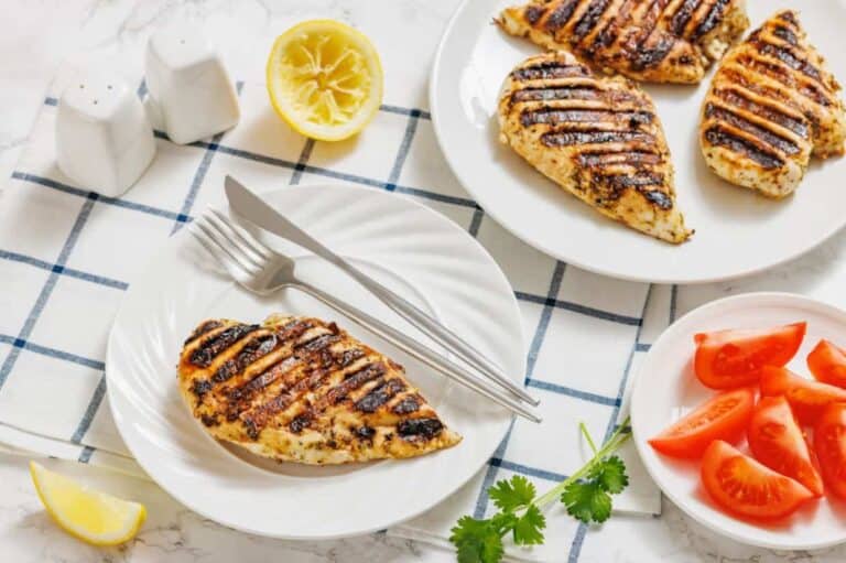 Grilled chicken breasts on plates, with sliced tomatoes, lemon halves, a sprig of cilantro, and salt and pepper shakers on a checkered cloth.