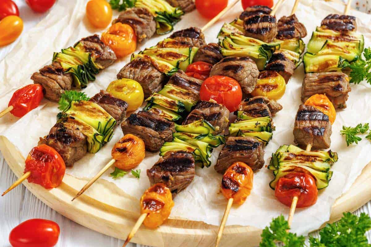 A wooden round serving board is covered with white parchment paper, displaying numerous grilled steak and veggie skewers arranged horizontally across it.