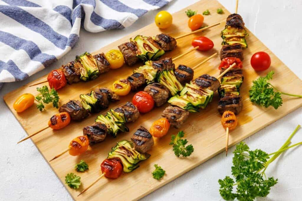 Grilled skewers with beef, cherry tomatoes, and zucchini on a wooden board, garnished with parsley, next to a striped white and blue towel.