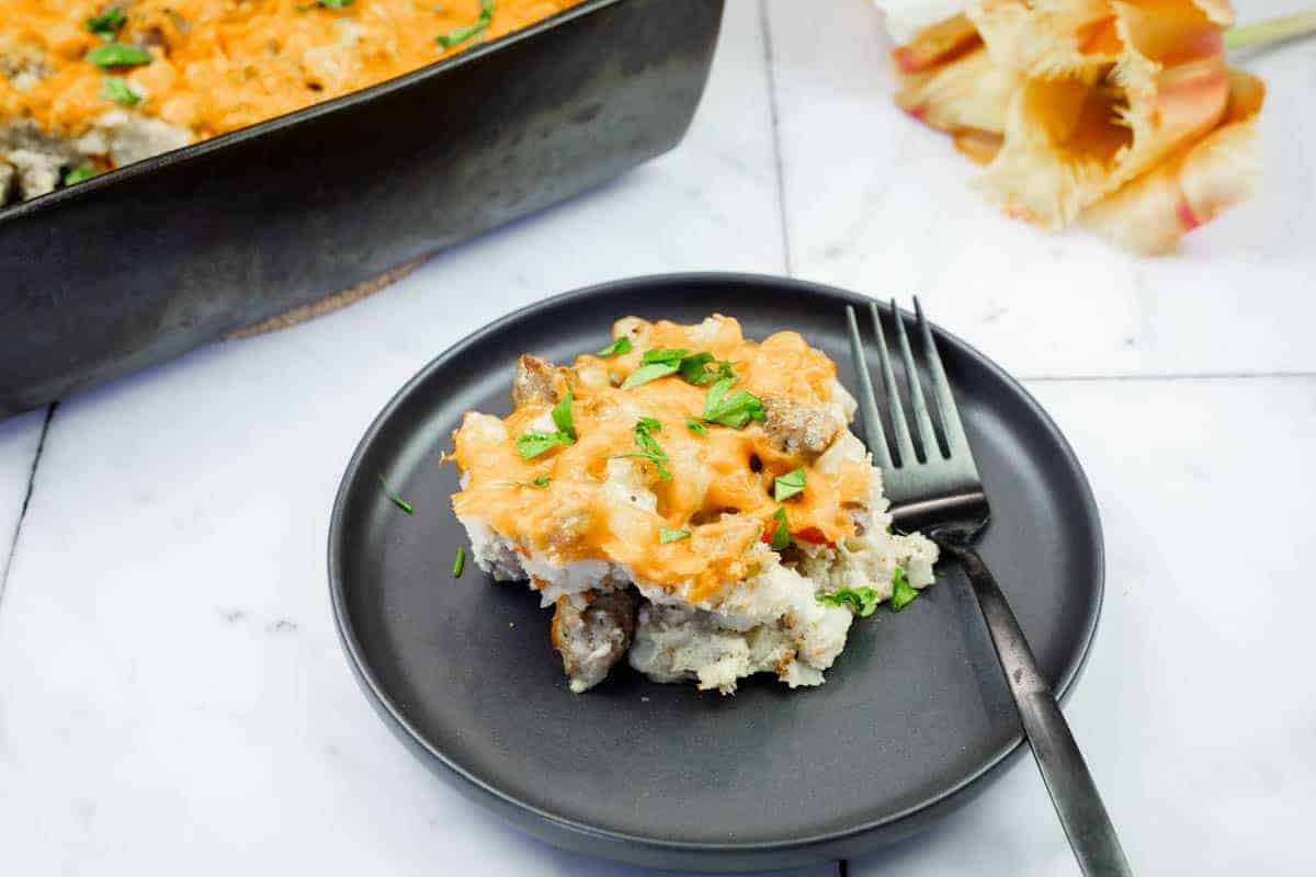 A serving of Sausage-Hash Brown Breakfast Casserole on a black plate with a fork, next to a baking dish and pasta shells on a marble surface.