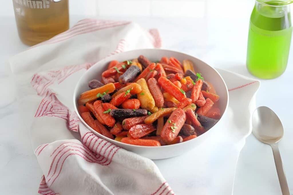 A white bowl filled with roasted baby carrots in various colors, garnished with herbs, sits on a red-striped cloth with a spoon and green drink nearby.