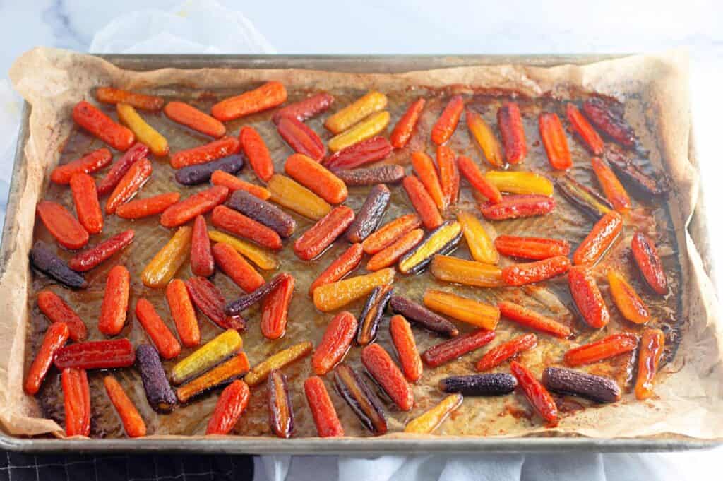 A baking sheet lined with parchment paper holds roasted baby carrots in red, orange, yellow, and purple varieties.